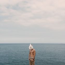 Man in dog mask standing by sea against sky