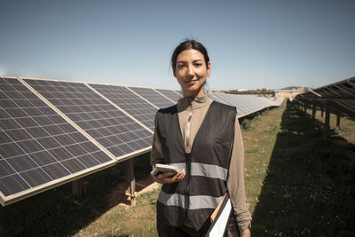 Portrait of smiling female maintenance engineer holding smart phone while standing near solar panels in field