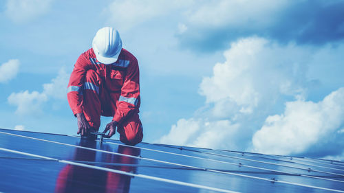Low angle view of male technician repairing solar panels against cloudy sky