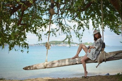 Woman sitting on log swing at beach against sea