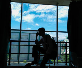 Silhouette man sitting by window against sky