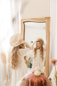 A beautiful teenage girl with long hair measures a straw hat in front of a mirror. self-admiration