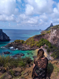 Rear view of woman looking at seascape while sitting on mountain against sky