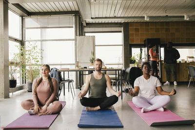 Full length of multiracial professionals meditating together on mats at office