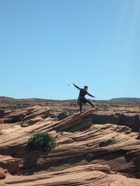 Rear view of man jumping on rock formations against clear sky