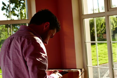 Rear view of man using mobile phone while standing against window