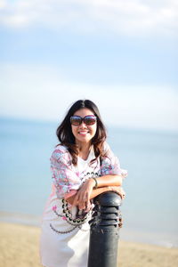 Portrait of young woman wearing sunglasses standing at beach against sky