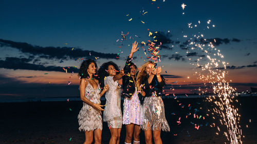 Cheerful young female friends with firework and confetti at beach against sky during sunset