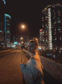 Side view of young woman standing on street against illuminated buildings