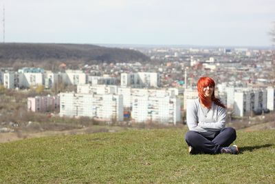 Portrait of young woman sitting on grassy field against cityscape