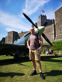 Portrait of mid adult man standing by airplane on grassy field against fort during sunny day