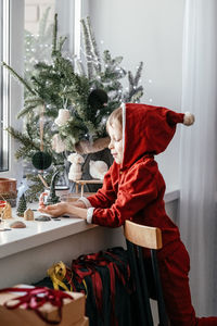 A thoughtful boy in a santa costume sits at the window and makes christmas decorations