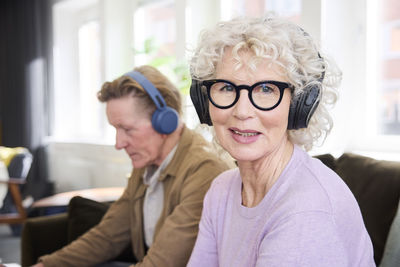 Smiling senior woman looking at camera while wearing headphones, senior man in background