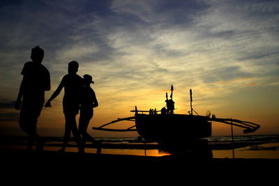 Silhouette men walking at beach with outrigger canoe in background during sunset