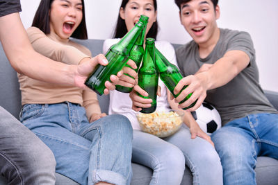 Friends toasting drinks while watching sports on sofa against wall