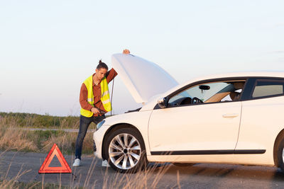 Man looking a broken car on the side of the road in an empty landscape with open car hood 