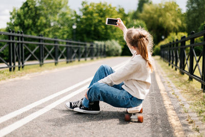 Cute blonde teenage girl sitting on a skateboard in a city park chatting on a smartphone 