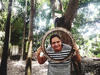 Portrait of smiling young woman peeking through old metal while standing against tree trunk