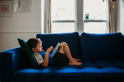 Young boy laying on blue couch playing educational games on tablet