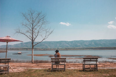 Rear view of young woman sitting on bench by lake against sky