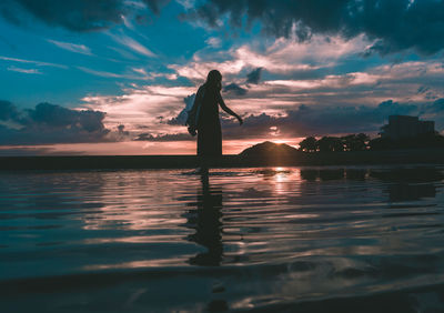 Silhouette woman standing at beach against cloudy sky during sunset