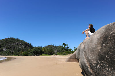 Low angle view of woman sitting on boulder at beach against clear blue sky