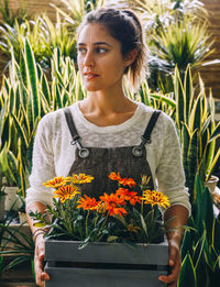 Young female owner of greenhouse holding in hands wooden tray with colorful margarita flowers while standing next to green tropical snake plants