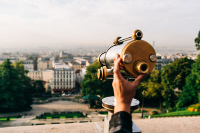Cropped image of hand holding telescope over cityscape against sky