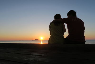 Rear view of couple sitting on shore against sunset sky