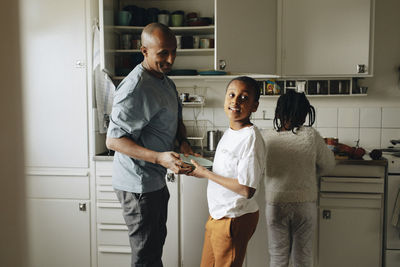 Son helping father while holding plates in kitchen at home