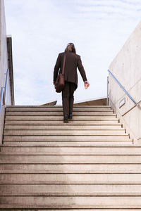 From below back view of unrecognizable stylish businessman in classy suit with briefcase and takeaway coffee climbing up stairs in city