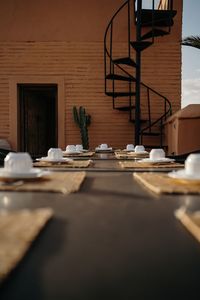 Sunlit terrace of modern restaurant with long table served with tea sets against terracotta wall with spiral staircase