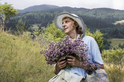 Portrait of smiling woman standing on field and holding oregano herbs flowers
