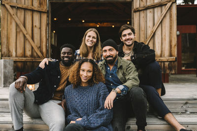 Portrait of smiling male and female friends sitting on staircase against cottage during social gathering