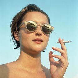 Portrait of woman wearing sunglasses while smoking cigarette against clear sky
