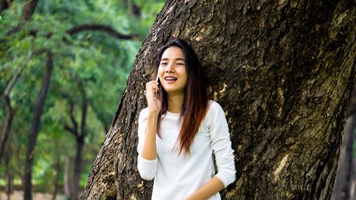 Smiling beautiful young woman talking on mobile phone while standing against tree