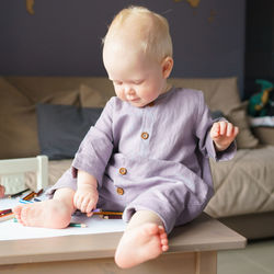 Cute blue eyed baby boy with golden hair sitting on table and playing with colored pencils