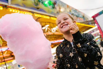 From below delighted girl smiling and eating sweet candyfloss while standing at funfair