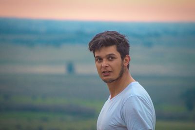 Handsome young man looking away while standing on landscape against sky during sunset