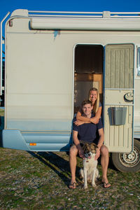 Young couple and a dog sitting at the entrance of a campervan.