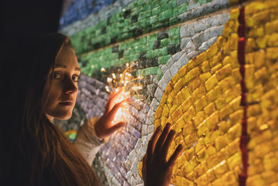 Close-up portrait of young woman with illuminated lighting equipment against wall
