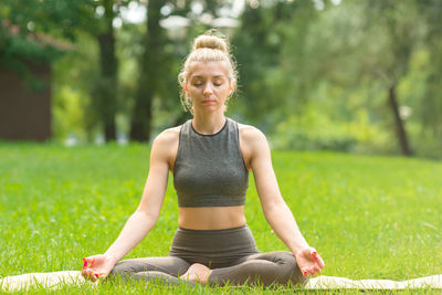 Slender woman sitting on the green grass in the park in sportswear and meditation