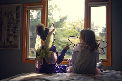 Rear view of girls in fairy costumes sitting on bed at home