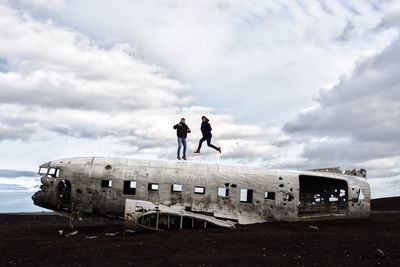 People jumping over abandoned airplane against cloudy sky