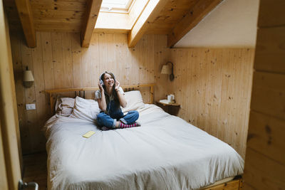 Smiling woman wearing wireless headphones sitting on bed