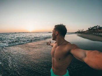 Shirtless man standing at beach against sky during sunset