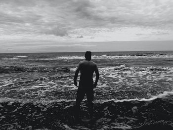 Rear view of man standing on shore at beach against cloudy sky