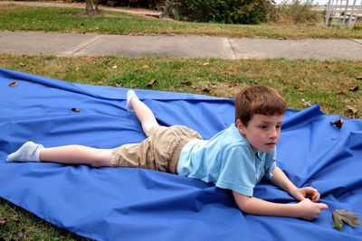High angle view of thoughtful boy lying on blue sheet in yard