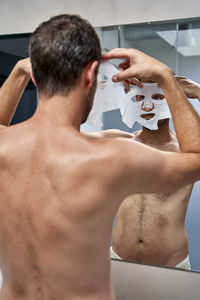 Man applying sheet mask on face in front of mirror at home