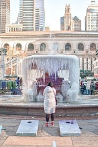 Rear view of woman standing against frozen fountain and built structure